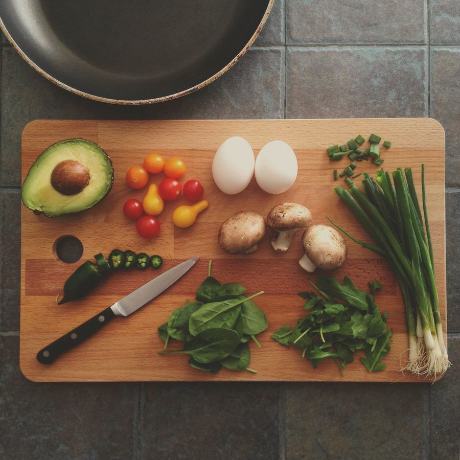 Ingredients on a wooden board before cooking: avocado, tomatoes, eggs, mushrooms, greens and a chef’s knife.